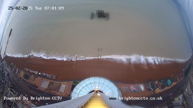 A bird's-eye view of a beach and ocean, taken from a webcam. The image shows a wide strip of brown sand with gentle waves lapping at the shore. In the water, a dilapidated structure is partially submerged, while the beach is lined with various objects and buildings. The sky above is cloudy, and time and date stamps are visible in the corner.