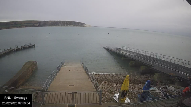 A coastal scene featuring a calm body of water with a cloudy sky overhead. In the foreground, there is a concrete ramp leading down to the water, flanked by a metal railing. To the left of the ramp, there are two wooden piers extending into the water, one with steps leading down. On the shoreline, a mix of pebbles and rocks is visible. There are several small boats and kayaks, one yellow, one blue, and one red, positioned on the right side near the railing. The distant background shows rocky cliffs and lush greenery. The time and date at the bottom indicate “25/02/2025 07:59:54.”