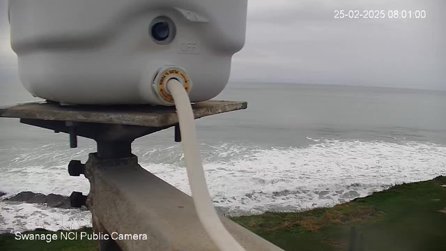 A close-up view of a white cylindrical object with a hose attached, sitting on a concrete ledge. The hose extends downwards towards the ocean below. The background features a gray, cloudy sky and rough waves crashing against the shore, with a hint of grass and land visible in the lower part of the image. The timestamp in the corner indicates the date and time.