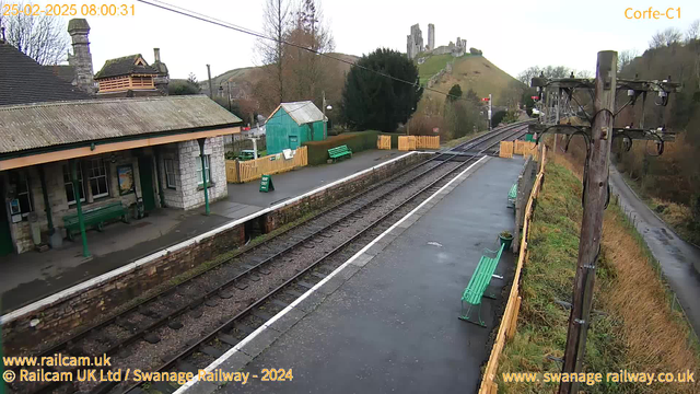 A view of a railway station during overcast weather. The foreground shows a platform with two green benches and a stone wall. To the left, there is a brick building with a peaked roof and windows. In the background, a hillside with a castle ruins is visible. The railway tracks run parallel to the platform and lead into the distance. There are trees and a wooden fence on the right side, along with a green sign indicating an exit.