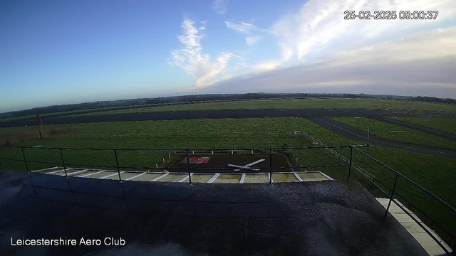 A view from a high vantage point overlooking an airport runway and grassy area. Below, there are white markings indicating the position of a small plane. To the left, a red flag is visible. The sky is mostly clear with some clouds, and distant green hills can be seen in the background. The image is timestamped with the date and time in the corner.