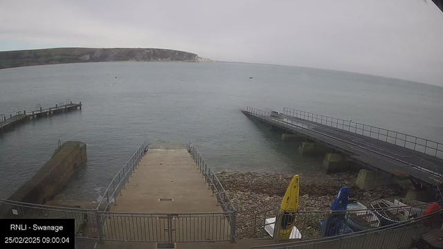 A coastal scene at Swanage showing a calm sea under a cloudy sky. In the foreground, a concrete ramp leads down to the water, flanked by railings. To the left, there is a wooden jetty extending into the water, and on the right, a wider jetty is visible. Several small boats are moored nearby, including two kayaks—one yellow and one blue. The rocky shoreline is visible alongside the ramp and jetties. In the distance, cliffs rise along the coast under a grey sky. The timestamp at the bottom shows the date and time as 25/02/2025, 09:00:04.