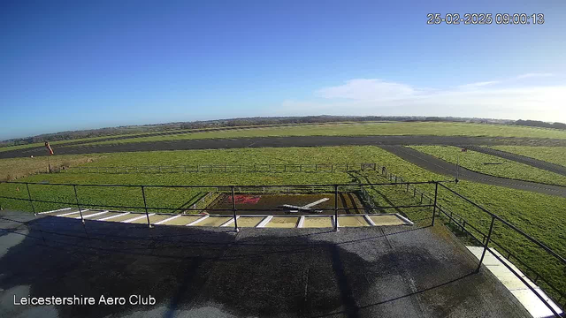 A clear blue sky is seen above a green landscape, with a large expanse of grass and some gravel areas. In the foreground, there is a raised platform with a railing, and a small model airplane is visible on the ground below. A windsock is located on the left, indicating wind direction. The landscape appears open, with a few fences in the distance, and the scene is well-lit, suggesting it is daytime. The date and time in the corner indicate it is 09:00 on February 25, 2025.