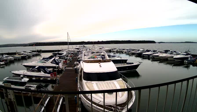 A marina filled with various boats and yachts is visible, tied up at docks. The water is calm and reflects the boats and surroundings. In the background, there are hills and a cloudy sky, with hints of blue peeking through. The scene conveys a serene atmosphere by the water.