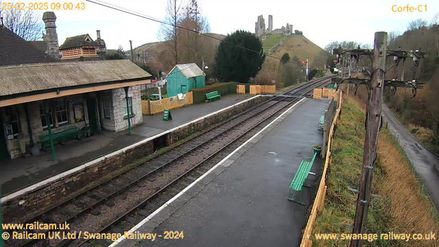 The image shows a railway station scene at Corfe, with a view of a stone building on the left featuring a peaked roof and a porch. Green benches line the platform, and there is a wooden fence with a sign that reads "WAY OUT." In the background, the landscape rises into a hill with the ruins of an ancient castle at the top. The tracks are visible on the right, leading off into the distance, with telegraph poles standing along the edge of the platform. The sky is partly cloudy and there are trees in the vicinity.