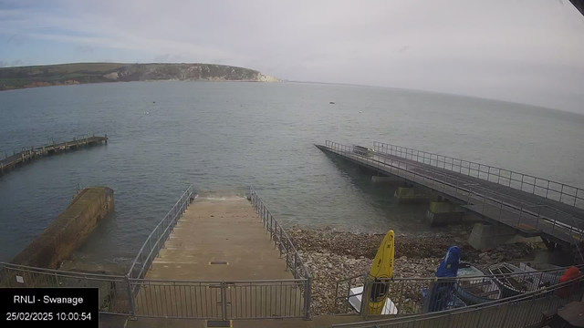 A view of a calm sea with a sandy shore in the foreground. To the left, there is a stone jetty extending into the water, and a wooden pier. Several boats are visible, including two yellow kayaks and one blue kayak, secured near the shore. In the background, a hilly landscape rises, leading to cliffs in the distance under a cloudy sky. The timestamp shows the date and time as 25th February 2025, 10:00:54.