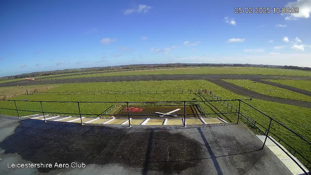 An aerial view from a rooftop at the Leicestershire Aero Club shows a wide expanse of green grass and runways. In the foreground, there is a light-colored airplane on the ground, with a red area nearby. The sky is bright blue with a few scattered clouds. The landscape features a fence along the edges of the field and a winding road in the background. The image is timestamped with the date and time in the corner.