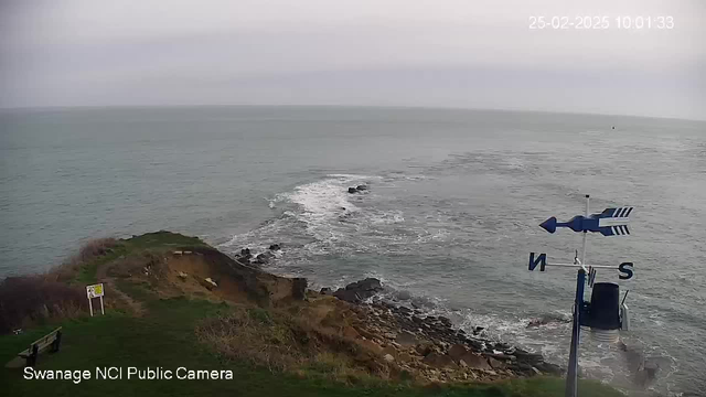 A coastal scene showing a rocky shoreline where waves are breaking. The water is a calm, muted blue, while the sky above is mostly overcast with gray clouds. In the foreground, there is a grassy area with a bench and a yellow sign, partially obscured by vegetation. To the right, there's a weather vane pointing northwest, indicating the cardinal directions. The date and time are displayed in the top right corner of the image.