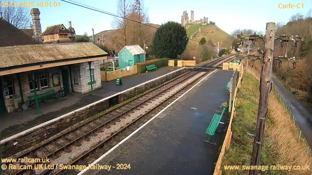 A railway station scene featuring a platform with tracks extending towards the right. In the foreground, there are several green benches along the platform. To the left, a building with a slate roof and stone walls displays large windows, while a small green shed is visible nearby. Beyond the platform, a hillside is dotted with trees, leading up to a castle-like ruin at the top. The sky is clear and blue.