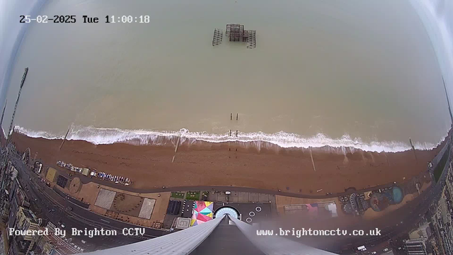 A high-angle view of a beach scene captured from a webcam. The image shows a sandy beach with gentle waves lapping at the shore. In the water, there is a partially submerged structure. The beach is bordered by a promenade, where colorful beach amenities are situated. There are sporadic people visible along the beach and walkway. The ocean appears grayish-blue, reflecting a cloudy sky.