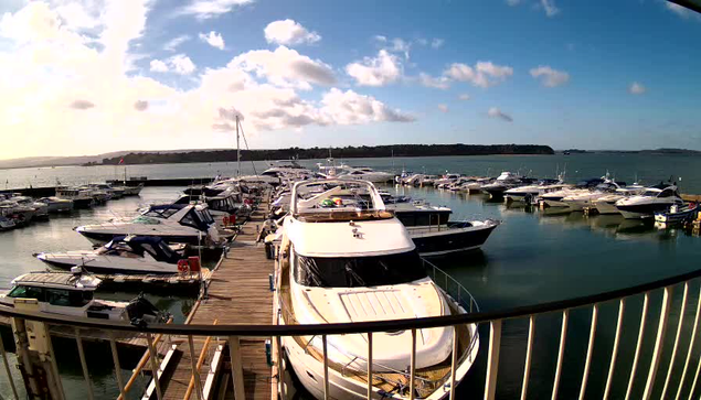 A marina filled with various boats and yachts docked along a wooden pier. The scene is set under a bright blue sky with scattered clouds, and the water is calm and reflective. To the background, there are green hills or land visible across the water.