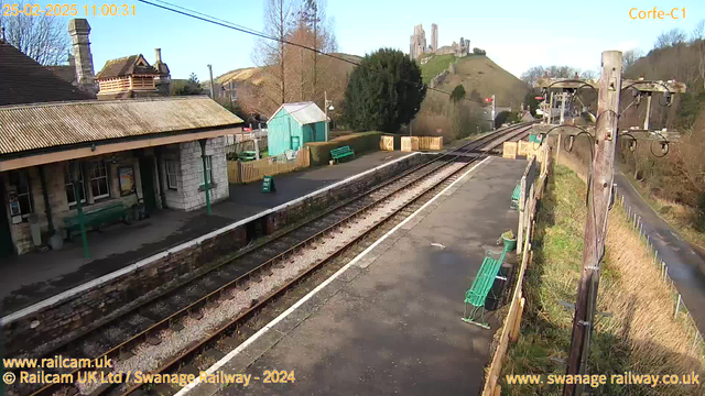 A railway station scene featuring a stone building with a peaked roof and multiple chimneys. To the left, there are two green benches positioned along a platform bordered by a stone wall. In the background, a hill rises sharply, topped by a castle ruin. On the right side of the image, a wooden pole supports electrical wires. The sky is clear and blue, and the station appears to be quiet, with no trains visible. A green shed is also present near the station building.