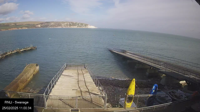 A view of a calm sea with a clear blue sky and a few scattered clouds. In the foreground, there is a wooden jetty extending into the water, with a low stone wall on the left side. There are two kayaks, one yellow and one blue, stored near the edge of the water on a rocky shoreline. In the background, cliffs can be seen along the coast, and a metal structure on the right extends further into the water. The image also includes a timestamp at the bottom left corner.