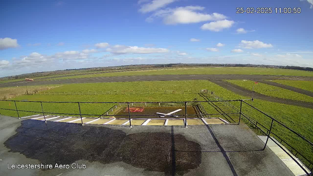 A clear blue sky with scattered white clouds occupies the upper half of the image. Below, a green grassy field stretches across the scene, with a small white model airplane placed on a landing pad marked in red. In the background, a tarmac runway is visible, and patches of dark wet ground indicate recent rainfall. A few fences are visible, outlining the field near the runway, which extends into the distance. The bottom left corner shows the name "Leicestershire Aero Club" in white text.