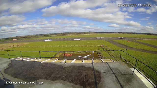 A view from a webcam showing an airfield on a sunny day. In the foreground, there is a section of a tarmac platform with markings. Behind it, several small airplanes are parked on a grassy field with some taxiway and runway markings visible. The background features a clear blue sky with fluffy white clouds and green hills in the distance. A windsock indicates the direction of the wind. The image captures a peaceful aviation environment.