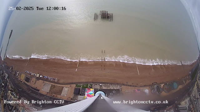 A high-angle view of a sandy beach meeting the ocean. The shoreline is visible with gentle waves lapping at the sand. A few wooden posts protrude from the water near the beach. In the background, an old pier structure is partially submerged. There is a colorful amusement area with a large, multi-colored tent on the beach. The image is timestamped at 12:00:16 on February 25, 2025.