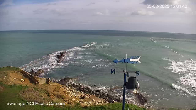 A coastal scene viewed from a webcam, showing a rocky shoreline with waves gently crashing against the rocks. The water is a mix of blue and green hues, stretching to the horizon under a partly cloudy sky. In the foreground, there is a weather vane on a pole with directional markings indicating north and south, along with a cylindrical structure below it. The landscape features grassy patches and rocky outcrops. The camera timestamp indicates the date and time as February 25, 2025, at 12:01:40.