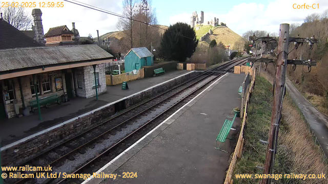 The image shows a railway station with a platform made of stone and concrete. There are several green benches along the platform. In the background, a small blue shed is visible, along with a fenced-off area containing more benches. To the right, a wooden pole with electrical wires is present. In the distance, a castle ruins sit atop a hill surrounded by trees. The sky is partly cloudy, creating a bright atmosphere.