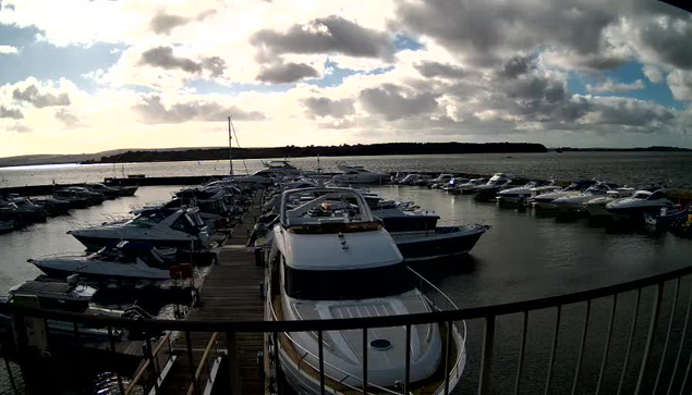 A view of a marina filled with various boats moored along a wooden dock. The scene features a large, white boat in the foreground, with several smaller boats lined up behind it. The water is calm, reflecting the cloudy sky above. The background reveals more boats and a distant shoreline with trees. The overall atmosphere is tranquil, with soft sunlight peeking through the clouds.