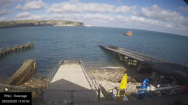 A coastal scene featuring a calm sea under a partly cloudy sky. In the foreground, there is a wooden pier extending into the water, with a light-colored ramp leading to the shore. On the right, two kayaks, one yellow and one blue, are resting on a rocky beach. In the background, a small orange boat is visible on the water, and a white cliff can be seen in the distance. The image captures a serene maritime environment with boats and the shoreline.