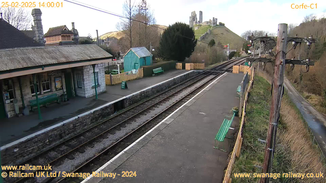 A railway station platform is shown with wooden benches and a small green shed in the background. The platform has a stone wall and a track running alongside it. In the distance, there is a hill topped with ruins of a castle. The sky is clear with some trees situated around the area. A sign on the platform reads “WAY OUT.” The scene is well-lit, indicating it is daytime.