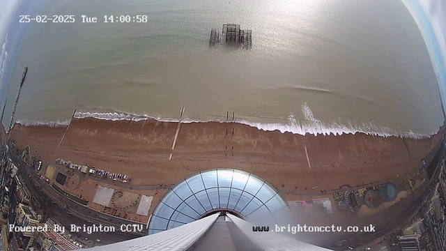 A high-angle view from a webcam overlooking a beach. The image shows a sandy shoreline with gentle waves lapping against it. In the distance, a pier extends into the water, and partially submerged remains of a structure are visible near the horizon. There are beachgoers and recreational areas along the coastline, including rides or attractions. The sky is partly cloudy, and a circular glass structure appears at the bottom of the frame, providing a view from above. A timestamp in the top left indicates the date and time.