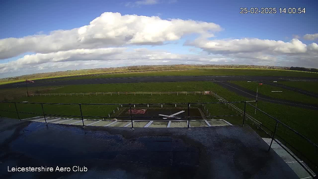 A view from a webcam at Leicestershire Aero Club shows a grassy airfield under a partly cloudy sky. The image features a large expanse of green grass bordered by a wooden fence on the left and a dark asphalt runway in the foreground. A windsock on a pole is visible, indicating wind direction. The scene captures the peaceful atmosphere of an airfield on a sunny day, with scattered clouds in the blue sky above.