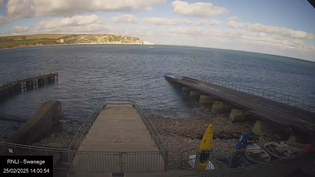 A coastal scene at Swanage featuring a calm sea under a partly cloudy sky. In the foreground, there are two piers extending into the water, one wooden and the other concrete with metal railings. To the right, a sandy area with small rocks leads to a kayak and other boats moored on the shore. The distant cliffs are visible in the background, with greenery on top. The time is noted as 14:00 on February 25, 2025.