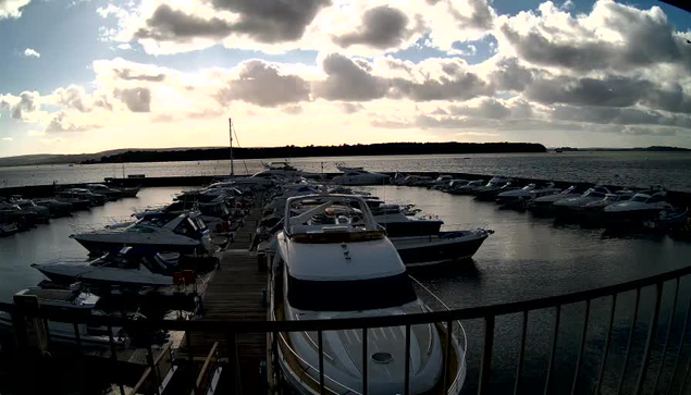 A marina is visible with numerous boats docked in the water. The scene is set against a cloudy sky, with soft sunlight filtering through the clouds. In the foreground, several white and blue boats are moored, while more boats are arranged further back in the marina. The water reflects the clouds and the light, creating a serene atmosphere. The horizon is lined with distant trees and land. A wooden dock is present on the left side of the image.