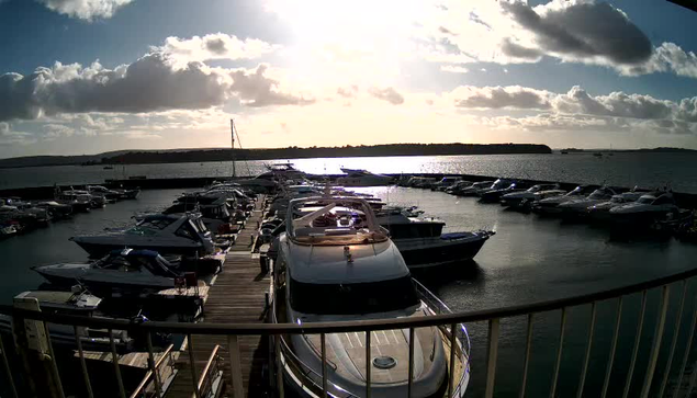 A marina filled with various boats moored closely together. The scene is illuminated by bright sunlight reflecting off the water, creating a sparkling effect. The sky is partially covered with fluffy clouds, and there are hints of land in the background. The perspective is from a higher vantage point, looking down towards the boats and water below.