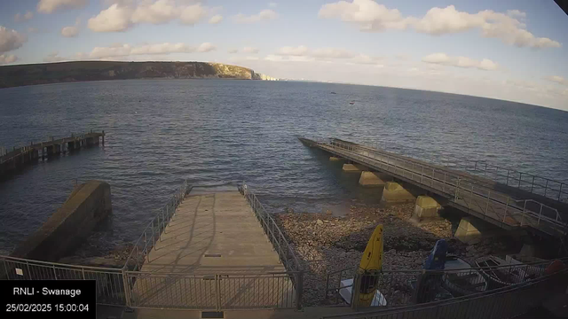 A view of a coastal area with calm water. In the foreground, there's a concrete platform leading to the water, with a railing along the edge. To the right, a sloped ramp extends into the sea, supported by pillars. There are some colorful kayaks parked on the shore, and pebbles cover the ground beneath them. The background features a steep cliff with greenery and white chalky areas, under a partly cloudy sky. The time and date are noted in the bottom left corner.