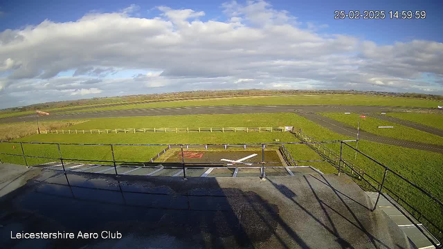 A wide view from a raised platform overlooking a green field and an airport runway. The ground is mostly grass, with some areas of asphalt. In the foreground, there is a white X marking the landing area. A wooden fence borders the field, and a red and white windsock is visible in the distance. The sky is partly cloudy with patches of blue.