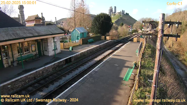 A view of Corfe Castle railway station captured by a webcam. The image shows a stone platform with a sloped roof, green benches, and a wooden fence. In the background, green hills rise steeply, featuring Corfe Castle at the top. The sky is clear with some clouds. Train tracks run along the platform, leading into the distance. There is a wooden shed painted blue and a sign indicating "WAY OUT" visible nearby.