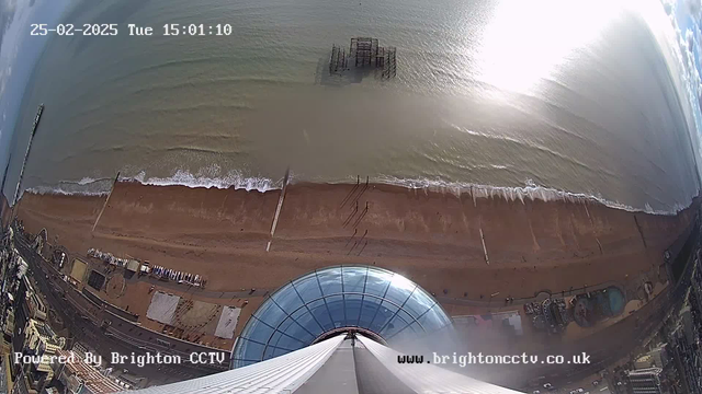 A high-angle view of a sandy beach with gentle waves lapping at the shore. At the center, there is a large, circular glass dome from a building, extending above the beach. In the background, a section of an old, partially submerged pier can be seen. The shoreline features a mix of sand and people, with some structures visible along the beach, including amusement rides. The sky is partly cloudy with bright sunlight reflecting off the water. A time and date stamp is displayed in the upper left corner.