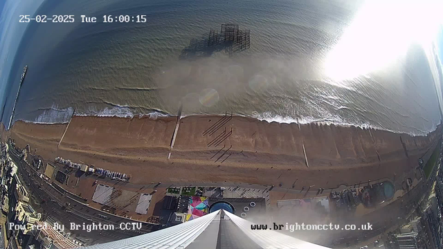 An aerial view of a beach on a sunny day, with gentle waves lapping at the sandy shore. The scene captures a stretch of beach lined with umbrellas and people walking. A pier extends into the water from the left side of the image, while remnants of an old pier structure can be seen partially submerged in the water. The coastline curves towards the horizon, and a few buildings are visible along the waterfront. The time stamp in the top left corner reads "25-02-2025, Tue 16:00:15."