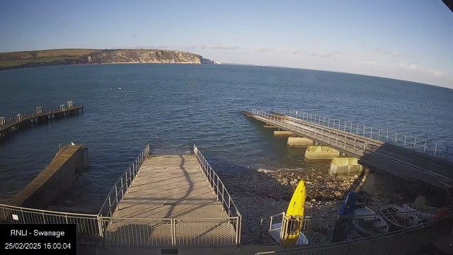 A seaside view showing a calm blue ocean under a clear sky. In the foreground, there are wooden piers extending into the water, with a yellow boat stored on the shore. To the right, there are concrete structures leading into the water. The shoreline has visible rocks and a few boats are scattered along the edge. In the background, green hills rise above the coastline, and cliffs are visible further out. The image has a timestamp indicating the date and time at the bottom left corner.