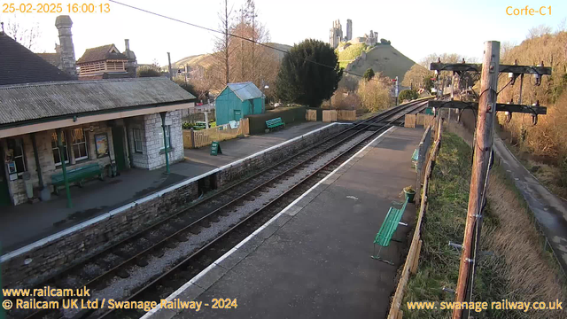 A view of a railway station platform during daylight. To the left, there is a stone building with a sloped roof, featuring large windows. A green bench is positioned on the platform in front of the building. In the background, a green hill rises, topped by a castle or ruin structure. Two wooden benches are visible on the platform, and a small blue shed can be seen further down the platform. A fence surrounds some grassy areas, and several railway tracks run through the scene. Overhead wires and a wooden utility pole are present on the right side. The image is timestamped with the date and time in the corner.