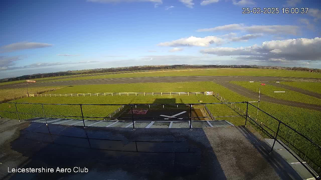 A wide view of an aerodrome under a clear blue sky with scattered clouds. In the foreground, a fenced area with a helicopter landing pad marked by a white cross. The grass surrounding the pad is vibrant green. To the right, an asphalt runway is visible, extending toward the horizon with various markings. In the background, soft hills are seen on the horizon, also covered in greenery. The time displayed in the corner indicates 16:00:37 on February 25, 2025.