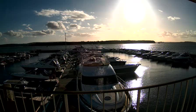 A marina scene during sunset with several boats docked in the water. The sun is low in the sky, casting a bright light that reflects on the water's surface, creating a shimmering effect. Light clouds are scattered across the sky, and the silhouettes of trees can be seen in the background along the shoreline. The setting provides a tranquil and picturesque atmosphere.