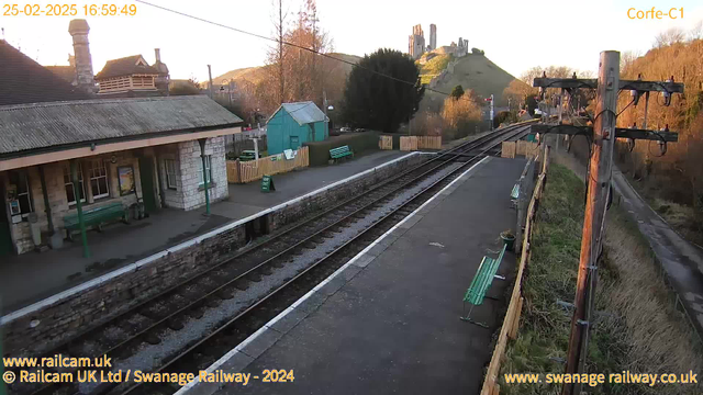 A railway station scene during sunset. In the foreground, there is a stone platform with several green benches and a waiting area. To the left, a stone building with a sloped roof features windows, while behind it, there is a small turquoise shed. The tracks are visible, leading into the distance. On the hill in the background, there are ruins of a stone castle. The surrounding area has trees and hedgerows, with a wooden fence marking the edge of the station property.