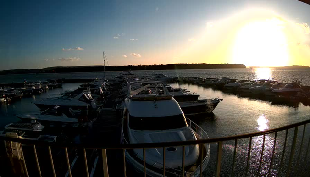 A marina filled with various boats is shown in the image, with several yachts moored in the water. The sun is setting in the background, casting a bright glow over the scene. The sky is mostly clear, with a few scattered clouds. The water reflects the colors of the sunset, creating a shimmering effect. The image is framed by a railing in the foreground.