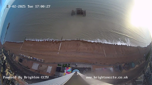 A high-angle view of a beach and the ocean taken from a webcam. The image shows the shoreline with sandy beach, gentle waves lapping at the edge of the water, and a pier extending into the sea. In the background, the ocean stretches out toward the horizon under a clear blue sky. There are scattered figures on the beach and some colorful structures visible at the edge of the sand. The time displayed in the corner indicates it's 5:00 PM on February 25, 2025.