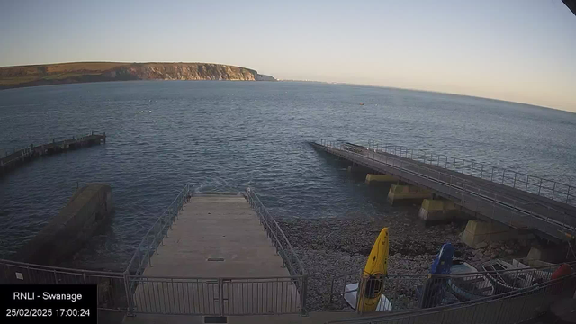 A scenic view of a coastline during sunset. In the foreground, a wooden jetty extends into the calm water. To the left, there is a rocky shoreline leading to the water, while several boats are visible, including a yellow kayak and a blue vessel. In the background, cliffs rise along the horizon, silhouetted against the sky. The water reflects the evening light, creating a tranquil atmosphere. The date and time displayed at the bottom indicate February 25, 2025, at 5:00 PM.