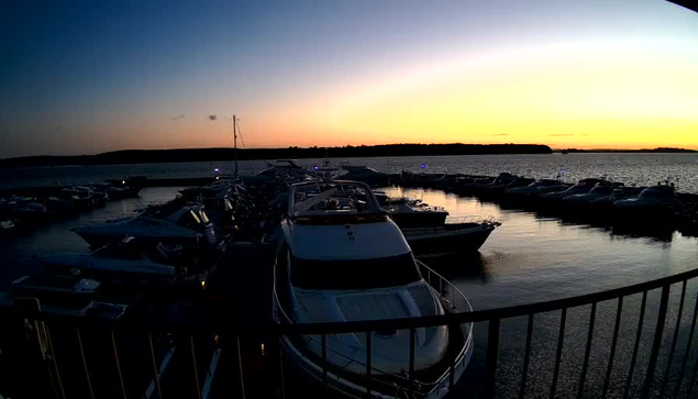 A tranquil marina scene at sunset, featuring several boats docked in calm waters. The sky transitions from soft orange to deep blue as the sun sets. Reflections of the boats and the changing sky are visible in the water, creating a peaceful atmosphere. A few lights on the boats are illuminated, adding a subtle glow to the scene. The horizon is marked by tree silhouettes.