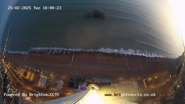 A high-angle view of a beach at sunset. The image shows the ocean with gentle waves, a sandy beach with some scattered objects, and a partially submerged pier in the water. The shoreline includes a colorful amusement area with a carousel. Illuminated buildings line the beach, and the scene is bathed in warm tones from the setting sun, creating a tranquil atmosphere. A timestamp in the corner indicates the date and time.