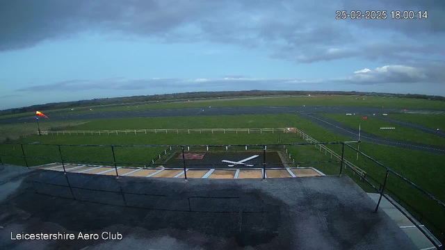 A wide-angle view of an aerodrome taken from a webcam. In the foreground, there is a dark rooftop with a white X marking an area, and a railing around the edge. Visible in the background is a green field, part of the aerodrome, intersected by dark runway paths. A windsock at the left is blowing in the wind, and various small aircraft are scattered on the ground. The sky is mostly clear with scattered clouds, and the scene is set in late afternoon light. The date and time are displayed in the top right corner.