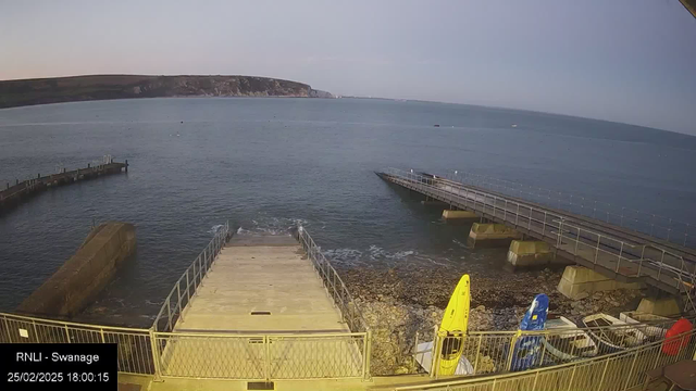 The image shows a coastal scene at a harbor during the evening. In the foreground, there are wooden ramps leading down from a metal platform to the water. Yellow and blue kayaks are parked near the edge of the platform on the right side. The water is calm, with a few ripples, reflecting the soft light of the setting sun. In the background, there are cliffs with greenery and a waterway extending into the horizon. The sky transitions from blue to soft purple as evening approaches.