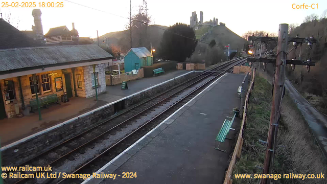 The image shows a railway station in the foreground with a stone building featuring a sloped roof and large windows. There are green benches on the platform, and a light-blue shed is visible nearby. In the background, a set of railway tracks runs towards the right side of the image, leading to a hilly area where a castle-like structure is situated on a hilltop. The sky is transitioning to twilight with warm hues, indicating the time is early evening. A power pole stands on the right side with electrical wires. The scene appears tranquil and rural.