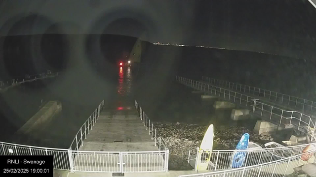 A dark scene depicting a pier by the water during nighttime. A small boat with lights is visible in the distance on the water, while the pier features a ramp leading down to the water's edge. There are two colored kayaks secured on the right side of the image, one yellow and one blue. The water appears slightly reflective from the boat's lights, and the shoreline is barely visible in the background. The image is relatively dark, with minimal lighting illuminating the area.