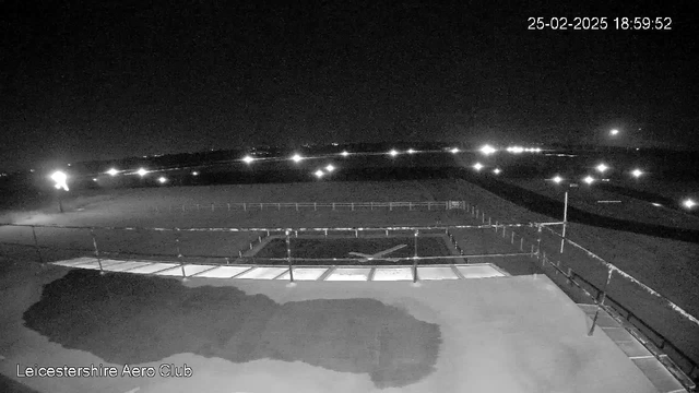 A black and white webcam image of an airfield at night. The foreground features a runway illuminated by distant lights from various sources. A dark area in the bottom left suggests a structure, possibly a building, but details are indistinct. In the background, there is a low fence marking the edge of the runway, with additional lights twinkling in the distance. The time and date are displayed at the top right corner of the image.